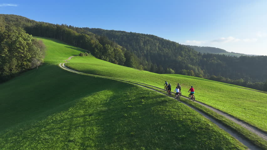AERIAL: Cycling friends enjoying a serene ride along a green hilltop bike trail bathed in gentle morning light with stunning views of rolling hills and nature. Mountain biking on a sunny autumn day.