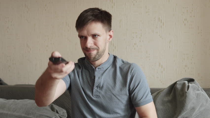 Caucasian man tries to press buttons on TV remote control while sitting on sofa. Non working device and negative emotions from unsuccessful attempts to turn on device with infrared remote control.