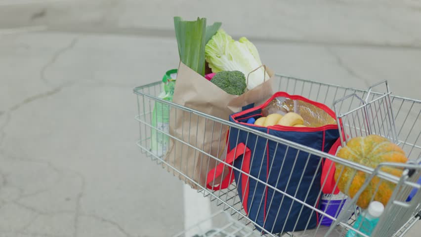 The detailed image captures a woman hands on the shopping cart handle, with the cart full of products in the background, set in the parking lot of a supermarket.