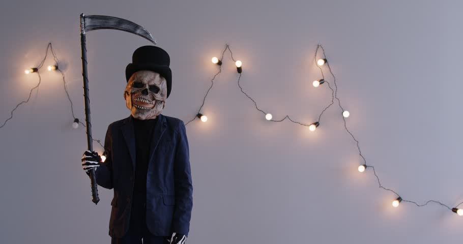 A boy in a scary mask and a black death suit holds a hand scythe, on a gray background. Halloween concept, mystery, symbol of death. Monster shakes his finger