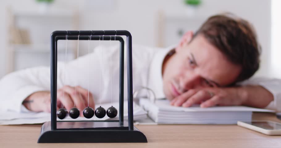 Bored businessman works with documents in his office, feeling burnout and tiredness. The presence of Newton Cradle on the desk highlights the monotony and boredom of the daily grind.