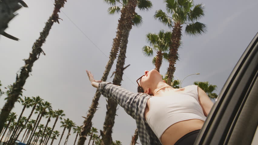 Young woman leans out of car window gracefully waving hand in rhythm of favorite music against palm trees. Cheerful girl in sunglasses enjoys summer holiday embracing journey by auto