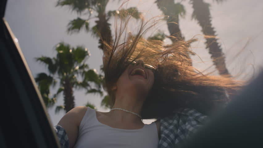 Excited woman enjoys the palm trees of seaside road. A girl leans out of window of rental car on a coastal highway on a sunny day. Hair in the wind. Tropical vacation on the beach of exotic islands
