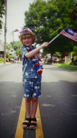 A young child joyfully waves a small US flag in the middle of a street, celebrating the fourth of July
