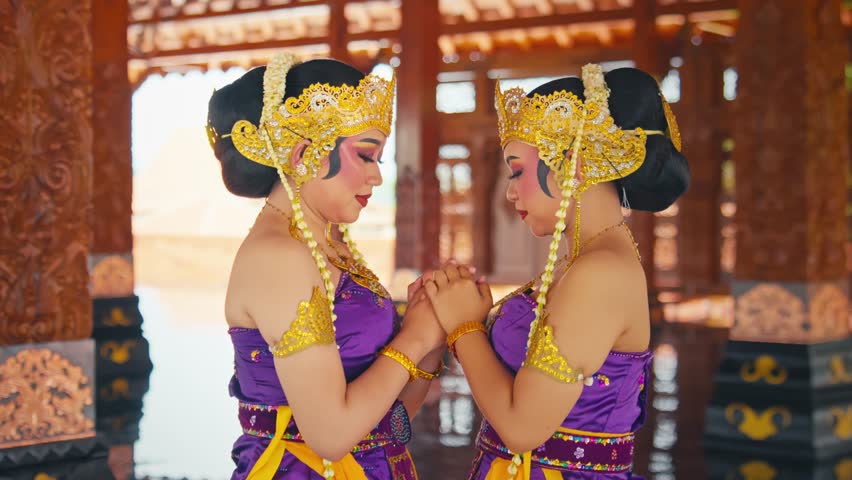 Two women in traditional Indonesian attire, wearing ornate golden headpieces and purple dresses, standing close together with their hands in a prayer position. The background features intricate wooden