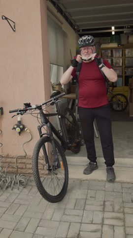 Vertical full shot of elderly male cyclist in helmet putting sunglasses on and walking away with bicycle while leaving garage and going for ride