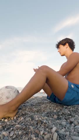 Wide shot hand held 4k vertical Video at 25 fps of a 19-Year-Old Young Man Sitting and Performing Soccer Tricks by Juggling a Ball on the Beach at Sunset