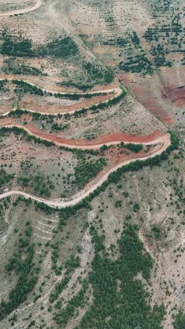 Pink truck navigates a long, winding mountain road, ascending through a stunning landscape of rugged terrain, valleys, and canyons under a clear blue sky