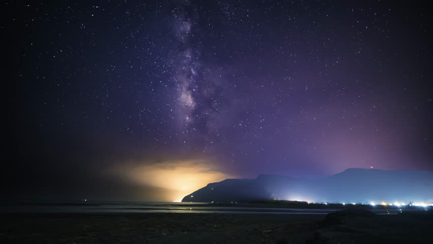 A serene night sky illuminates the coastal mountains. The Milky Way stretches across the horizon, casting a soft glow over the tranquil seascape. Suao, Yilan County, Taiwan.
