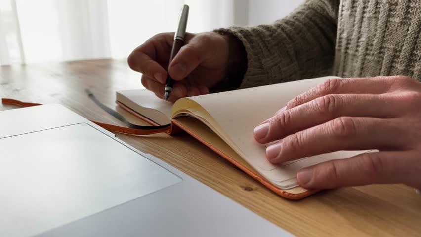 Male hand writing with a fountain pen in paper notebook on wooden table. Freelancer in beige wool sweater handwriting a note.