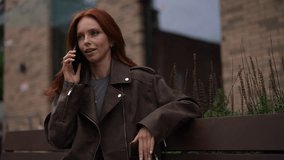 Cheerful young woman with long red hair wearing brown leather jacket sitting on bench, talking on phone and laughing. Happy redhead female talking on smartphone in city street. Shooting in slow motion - Powered by Shutterstock - Get 15% off with code: PIKWIZARD15