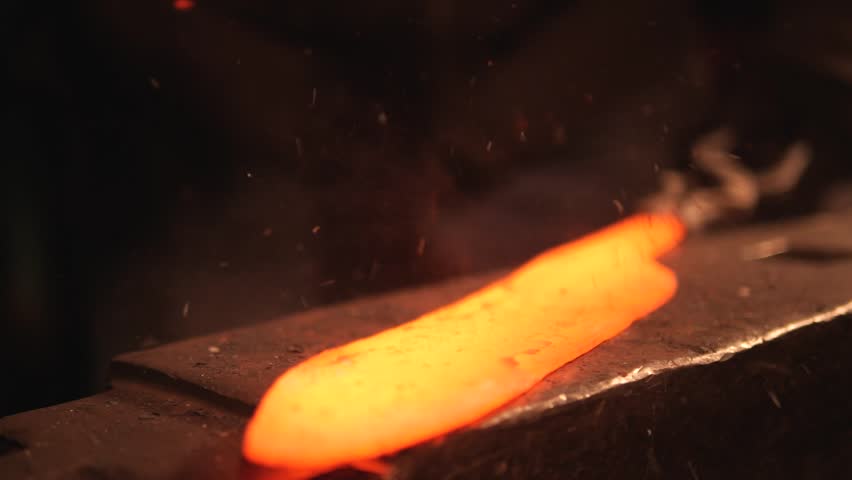 Working blacksmith shaping hot knife on anvil. Close up of male blacksmith hammering metalwork on anvil.  Red hot glowing ingot placed on an anvil and hit with blacksmith