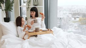 Mother and daughter sharing breakfast in bed, smiling at each other while holding croissants, bonding over a cozy morning. Wooden tray with croissants and cups on bed creates warm, intimate atmosphere - Powered by Shutterstock - Get 15% off with code: PIKWIZARD15