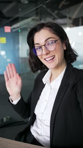 POV Smiling businesswoman waving at camera during video call in modern office. Professional woman engaging in virtual meeting using phone, showing friendly and communicative interaction Vertical video