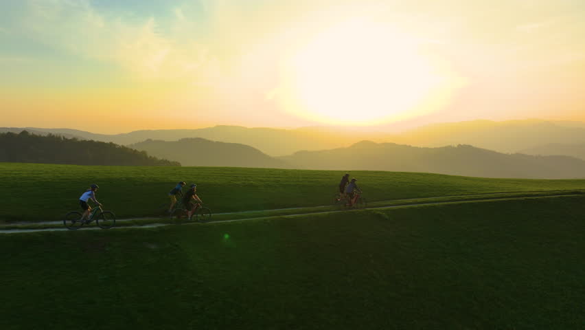 AERIAL, LENS FLARE: Mountain bike riders riding through rolling hills at golden sunset, with the warm glow of evening sky illuminating hilly landscape. Beauty and tranquillity of outdoor adventures.