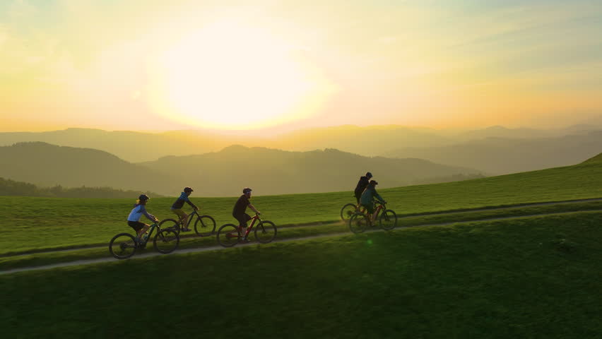 AERIAL, LENS FLARE: Mountain bike riders riding through rolling hills at golden sunset, with the warm glow of evening sky illuminating hilly landscape. Beauty and tranquillity of outdoor adventures.