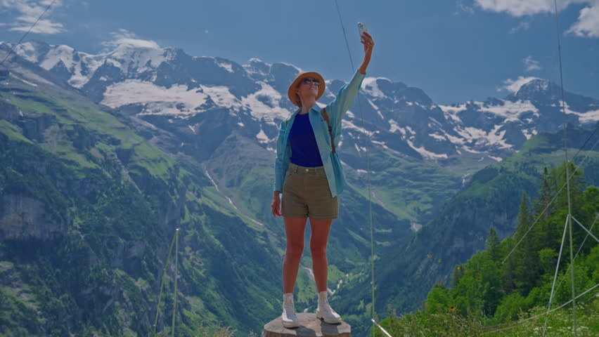 A girl in a hat in the mountains of Switzerland takes a selfie on her phone against the backdrop of beautiful alpine mountains.
