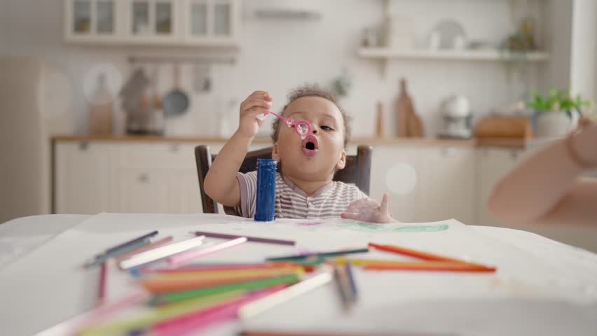Portrait of cute African little child sitting at table and blowing soap bubbles. Happy children party at home, kids have fun and joy, funny black toddler playing, slow motion, family and parenting