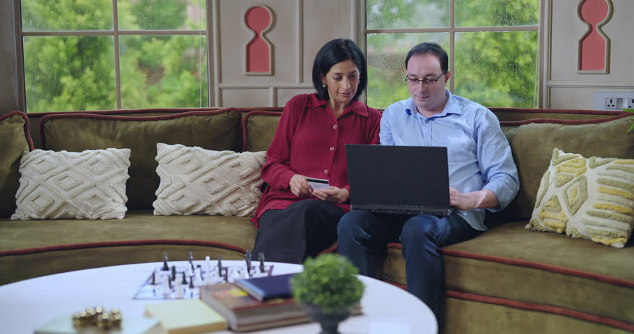 Indian happy old aged senior gen x couple sitting together cozy sofa use laptop look screen hold credit debit card do online shopping purchase goods buy furniture for new house enjoy day indoor home