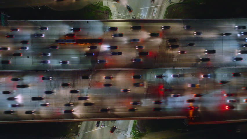 An aerial photo taken at night captures a crowded traffic jam in downtown Los Angeles. The freeway lanes are full of cars under the city lights, showcasing the bustling urban setting. Time lapse in 4K