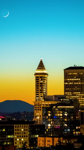 Time lapse of the Smith Tower in Seattle silhouetted against a vibrant dusk sky with a crescent moon setting above. The cityscape captures the calm transition from day to night.