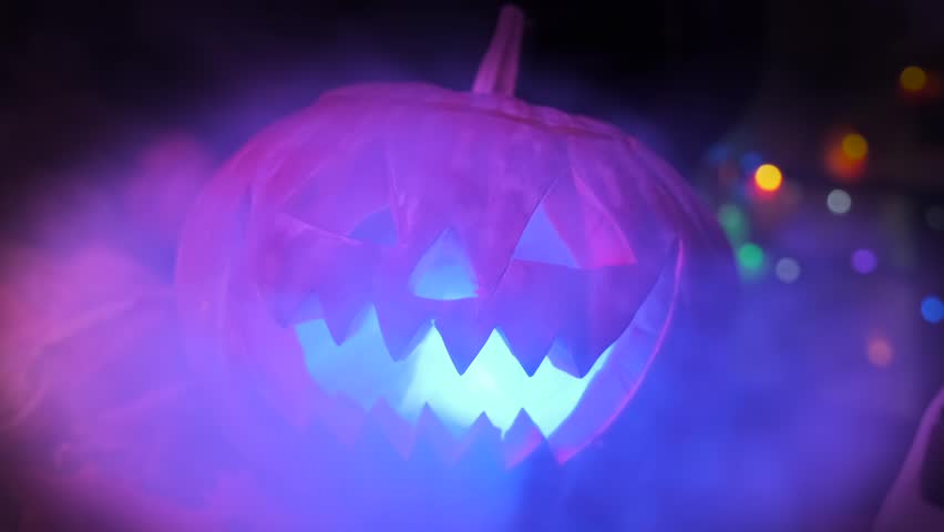 Close-up of witch hands caressing a carved spooky Halloween pumpkin in cold smoke next to a human skull with a burning candle and an apple among dry foliage.