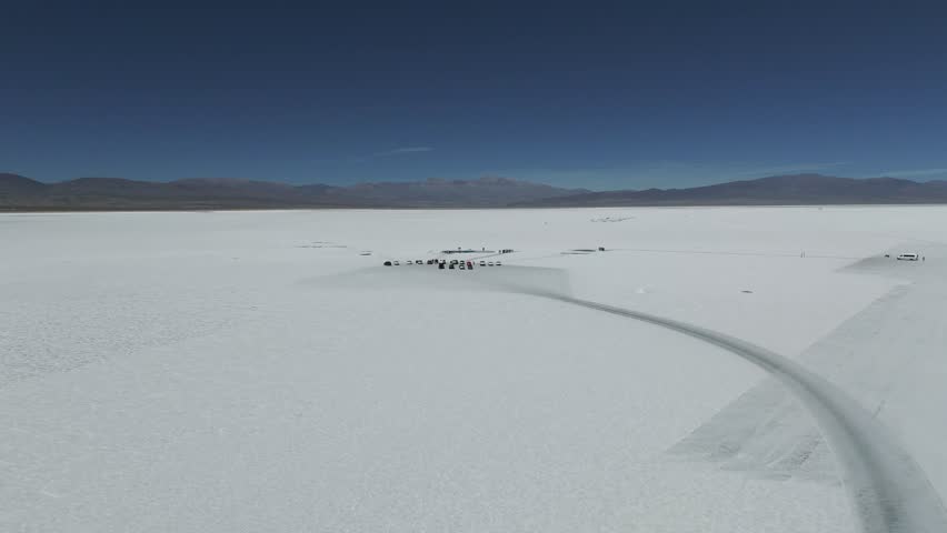 Drone aerial views nature scenes, a salt flat setting, an aerial view of Salinas Grandes, vehicles and people gathered near pools on a vast white area with mountains in the distance, pan forward