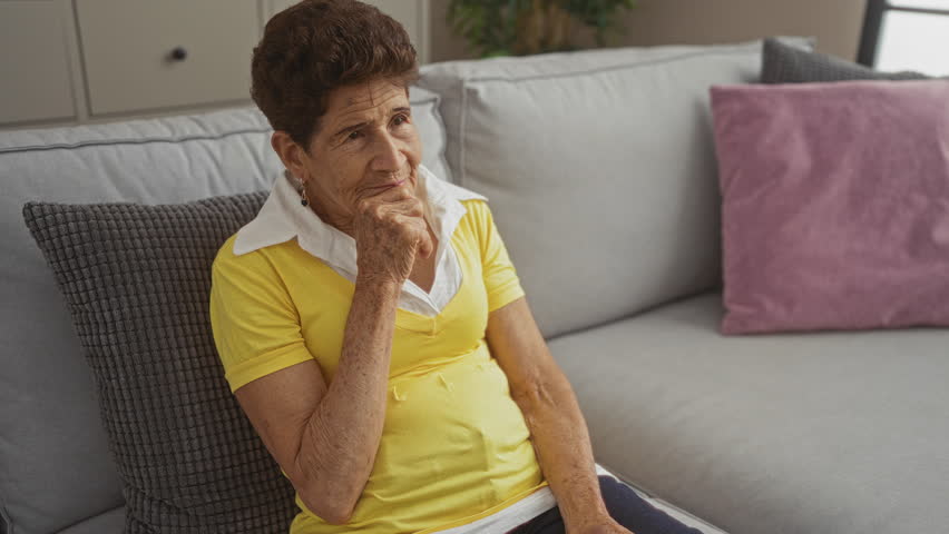 Elderly woman with short hair sitting in a living room, wearing a yellow shirt, holding a tv remote, and looking thoughtful while seated on a gray couch with colored pillows around.