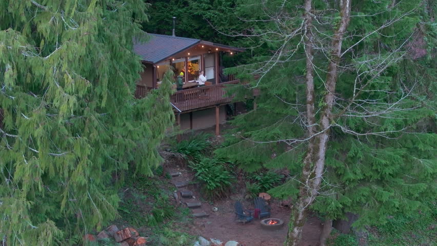 A wooden cabin in the forest near a river, with a fire pit burning nearby. A young couple stands on the balcony enjoying the view. Washington. USA. Aerial. 4K footage.