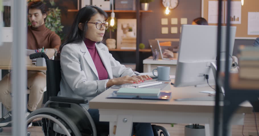 Female office worker disabled woman working with computer typing sitting in wheelchair at desk. Business and inclusive workspace concept.