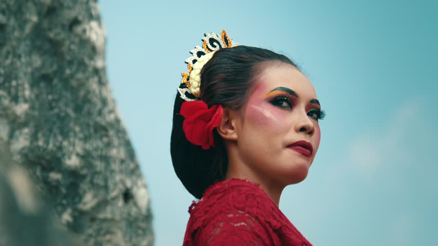 A woman in traditional attire with vibrant makeup, including colorful eyeshadow and a red flower in her hair. She stands against a scenic backdrop, exuding confidence and grace.