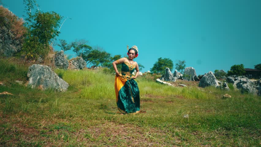A dancer in traditional attire performs on a grassy hillside surrounded by rocks and trees. The dancer is kneeling, with one hand raised and the other extended forward, showcasing a graceful movement