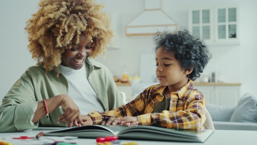 Little African American boy of preschool age sitting at table in the living room, watching photo book and discussing pictures with happy mother
