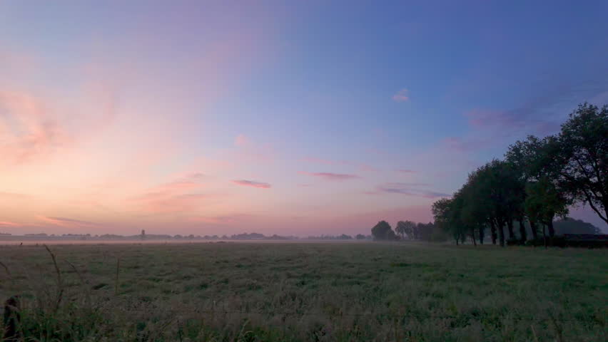 A peaceful and serene sunrise beautifully casts enchanting colors all over a misty, lush green field filled with trees