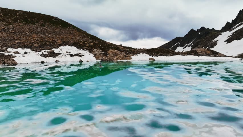 A man swims on a paddleboard in a mountain turquoise lake partially covered with snow, surrounded by snow-capped mountain peaks. Aerial view