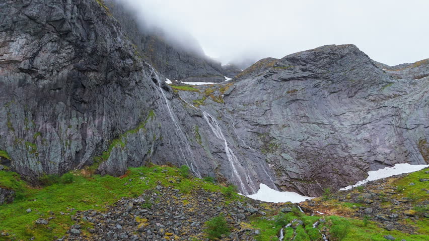 Ominous low angle fixed shot left of a mountain range in Vassdalsvattnet near Reine in Lofoten Norway. Waterfall is gushing down a rock face towards snow. Sky is cloudy, overcast and dark.