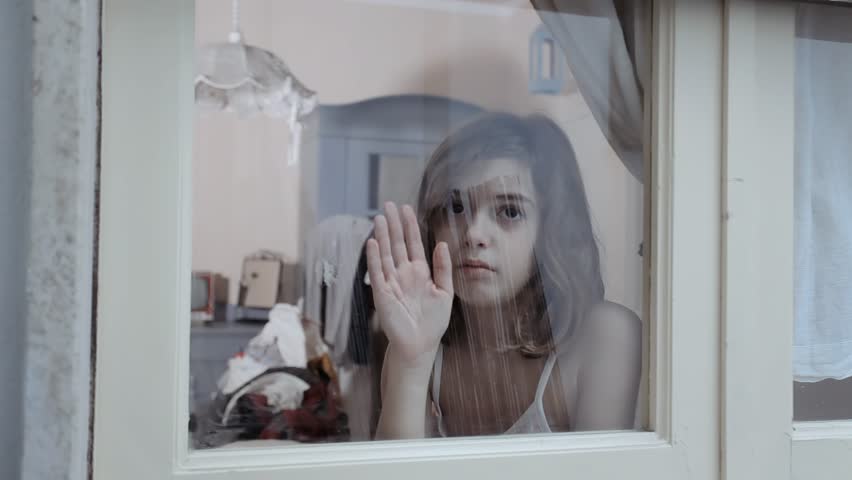 Young girl presses hand against window, gazing out with melancholic expression. Soft focus interior backdrop. Evokes feelings of loneliness, confinement, and yearning for connection.