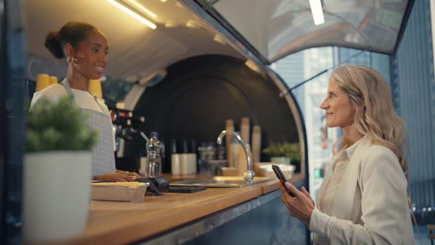 Friendly African American Food Truck Barista Serving Sparkling Water to a Businesswoman at a Modern City Location. Satisfied Customer Using a NFC Mobile Payment Application to Pay for the Order - Powered by Shutterstock - Get 15% off with code: PIKWIZARD15
