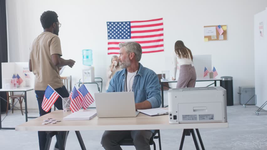 Handsome member of polling station kindly smiling to voters. Participating in elections. Man sitting at laptop in office or poll station. Gifting badges with American symbols to citizens.