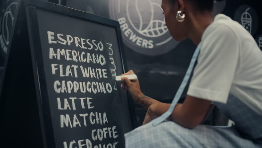 Black Female Food Truck Worker in an Apron Writes a Coffee Menu on a Chalkboard, Listing Drinks Like Espresso, Cappuccino, Latte, Matcha. Writing Down Prices and Preparing to Welcome Clients