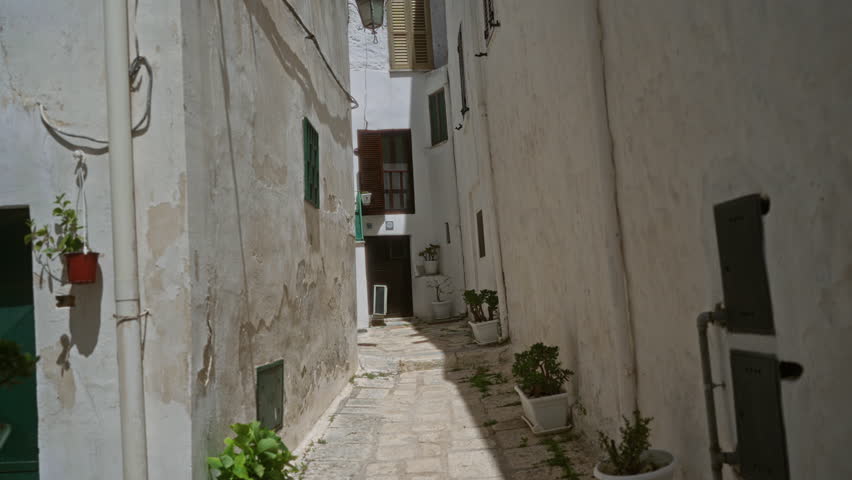 Charming narrow alley in ostuni, puglia, italy with whitewashed walls and potted plants, bathed in sunlight highlighting the picturesque street scene.