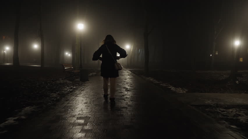Silhouette of a woman running along a park alley at night with streetlights shining through the fog