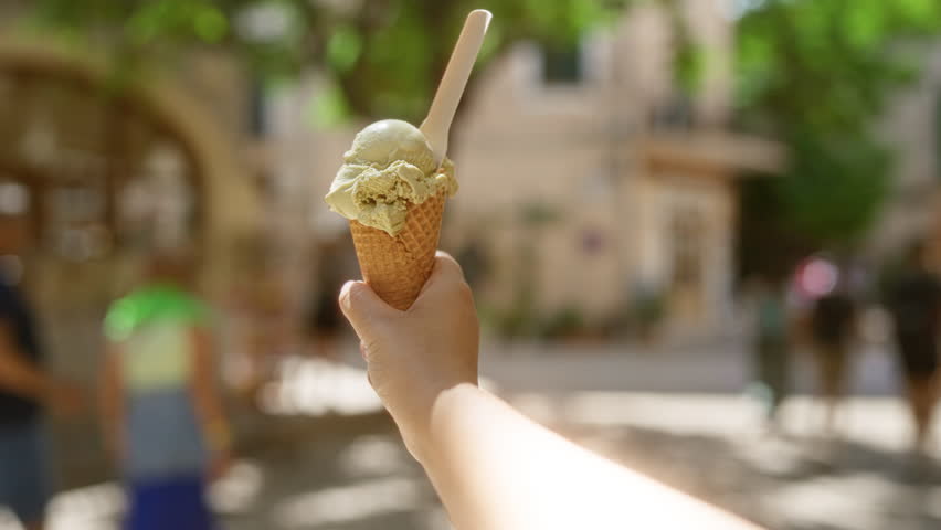 Hand holding an ice cream cone with trees and blurred people in the background on a sunny day outdoors
