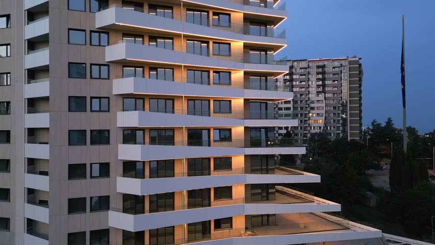 Aerial drone shot of a modern building facade illuminated by sleek LED lighting at dusk, capturing the contrast between the urban night skyline and contemporary architecture