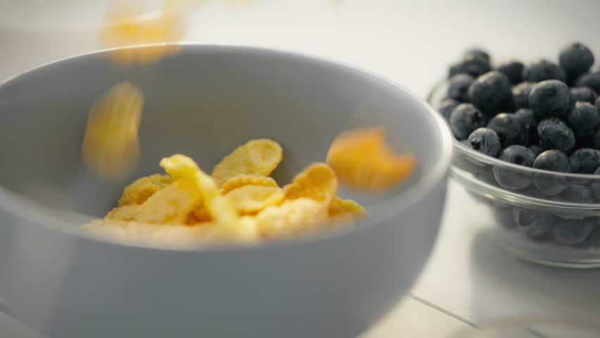 A slow-motion video shows cereal flakes slowly falling into a bowl. The bowl is filled with cornflakes, which are being prepared for a healthy person's breakfast