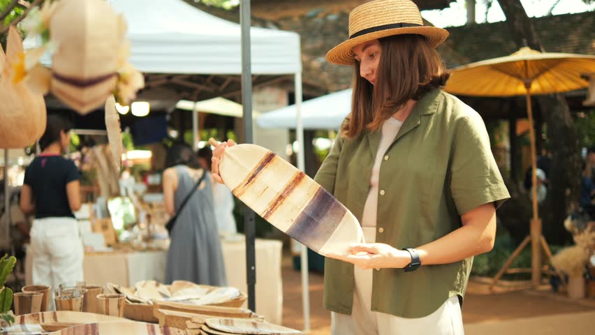 Pleased Girl in a Linen Green Shirt and Straw Hat Picks Out Handmade Plates at the Weekend Jing Jai Market, Having Fun. Vacation, Asian Culture, Shopping Concept