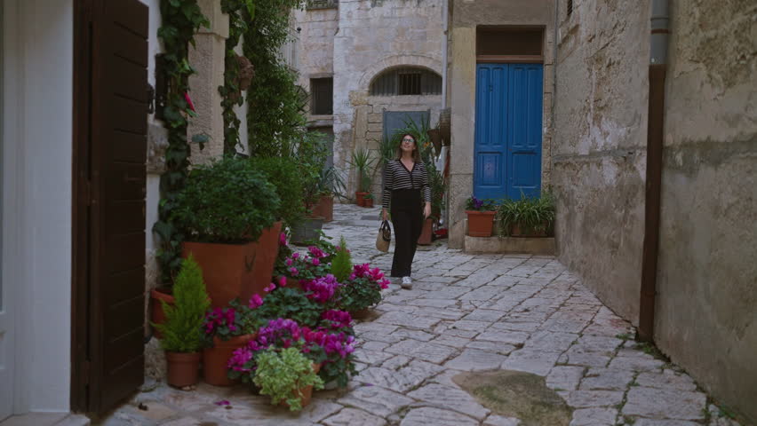 A young hispanic woman strolls through the picturesque streets of polignano a mare in puglia, italy, surrounded by vibrant flowers and rustic architecture.