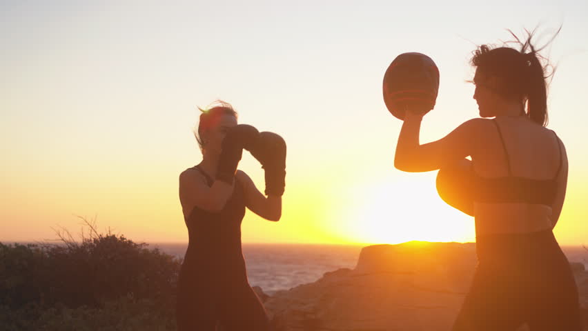 Determined backlit female fighters practicing defence boxing at beach during bright sunset. Strong young women boxers are exercising in nature. 