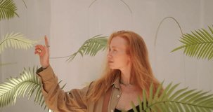 Portrait of a young red haired woman at an outdoor contemporary interior design exhibition. She stands inside the exhibit, with hanging paper palm leaves floating around her. She looks around. - Powered by Shutterstock - Get 15% off with code: PIKWIZARD15