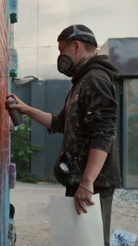Medium full vertical side shot of young male graffiti artists in gas mask, stained hoodie busy working on large colourful mural on brick wall in city street or yard
